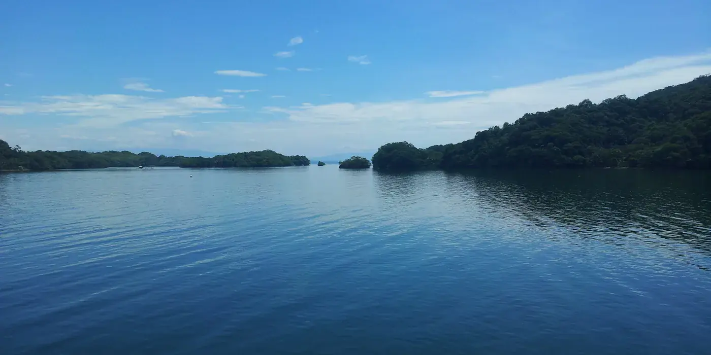 Flat, glassy water in the Gulf of Nicoya on the approach to Paquera, with low forested headlands closing in on both sides under a blue sky