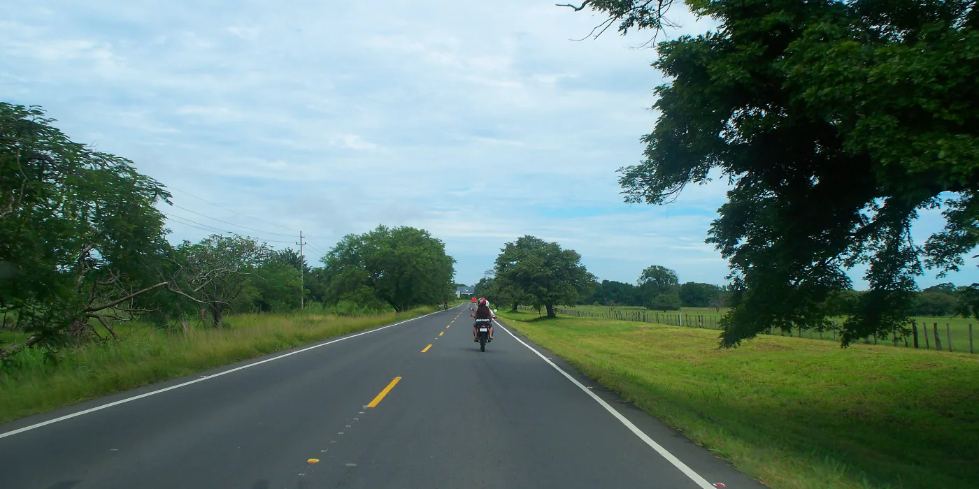 Driver’s view along the Pan-American Highway in Guanacaste, Costa Rica: a two-lane road with a yellow centre line running between green pasture and guanacaste trees, a motorcyclist ahead in the distance