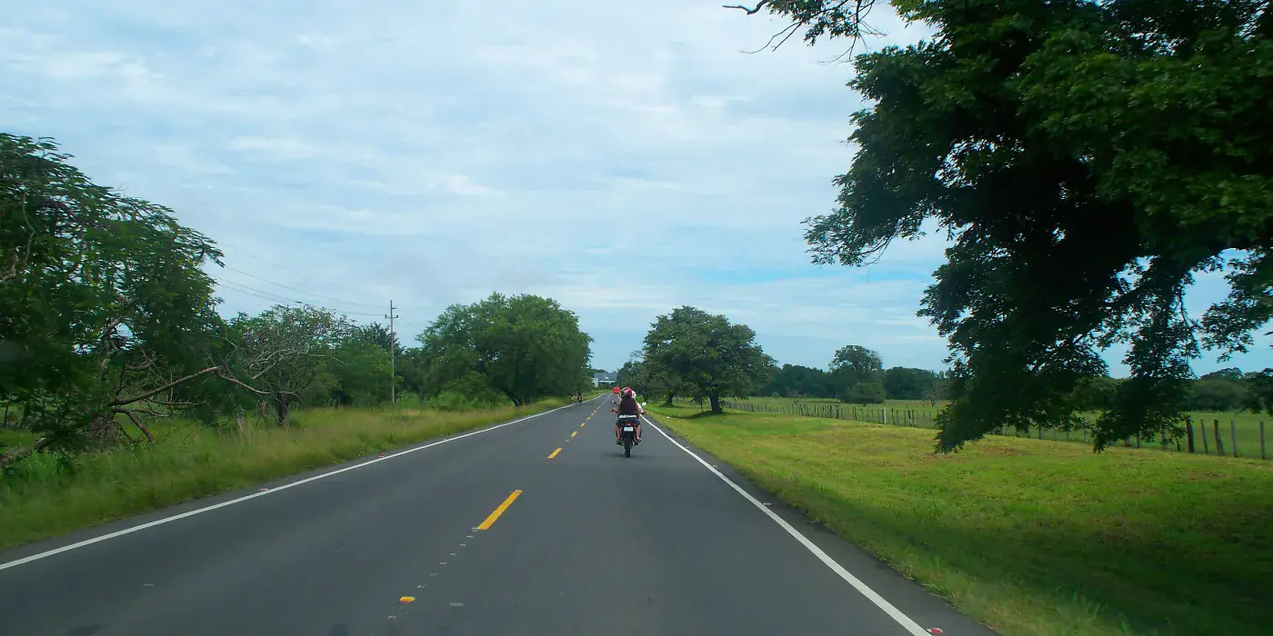 Driver’s view along the Pan-American Highway in Guanacaste, Costa Rica: a two-lane road with a yellow centre line running between green pasture and guanacaste trees, a motorcyclist ahead in the distance