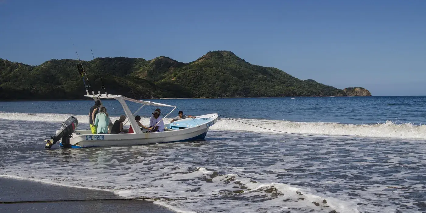 A small open boat loading passengers in the shallows off a Guanacaste beach, with forested headlands behind it under a clear sky