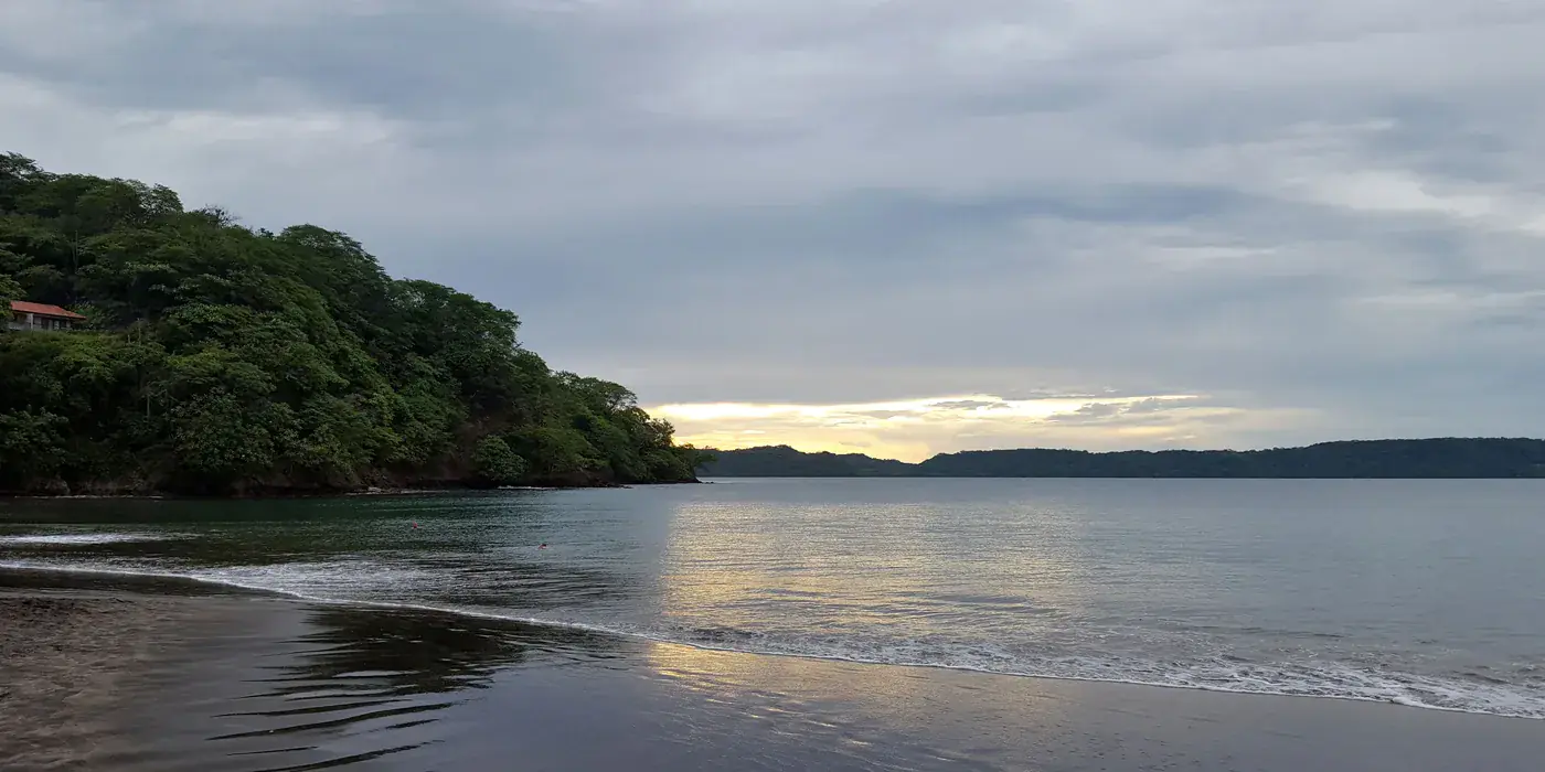 Bahía Culebra at dusk: a sheltered bay with flat water, a forested headland on the left and a thin line of gentle surf running up dark sand