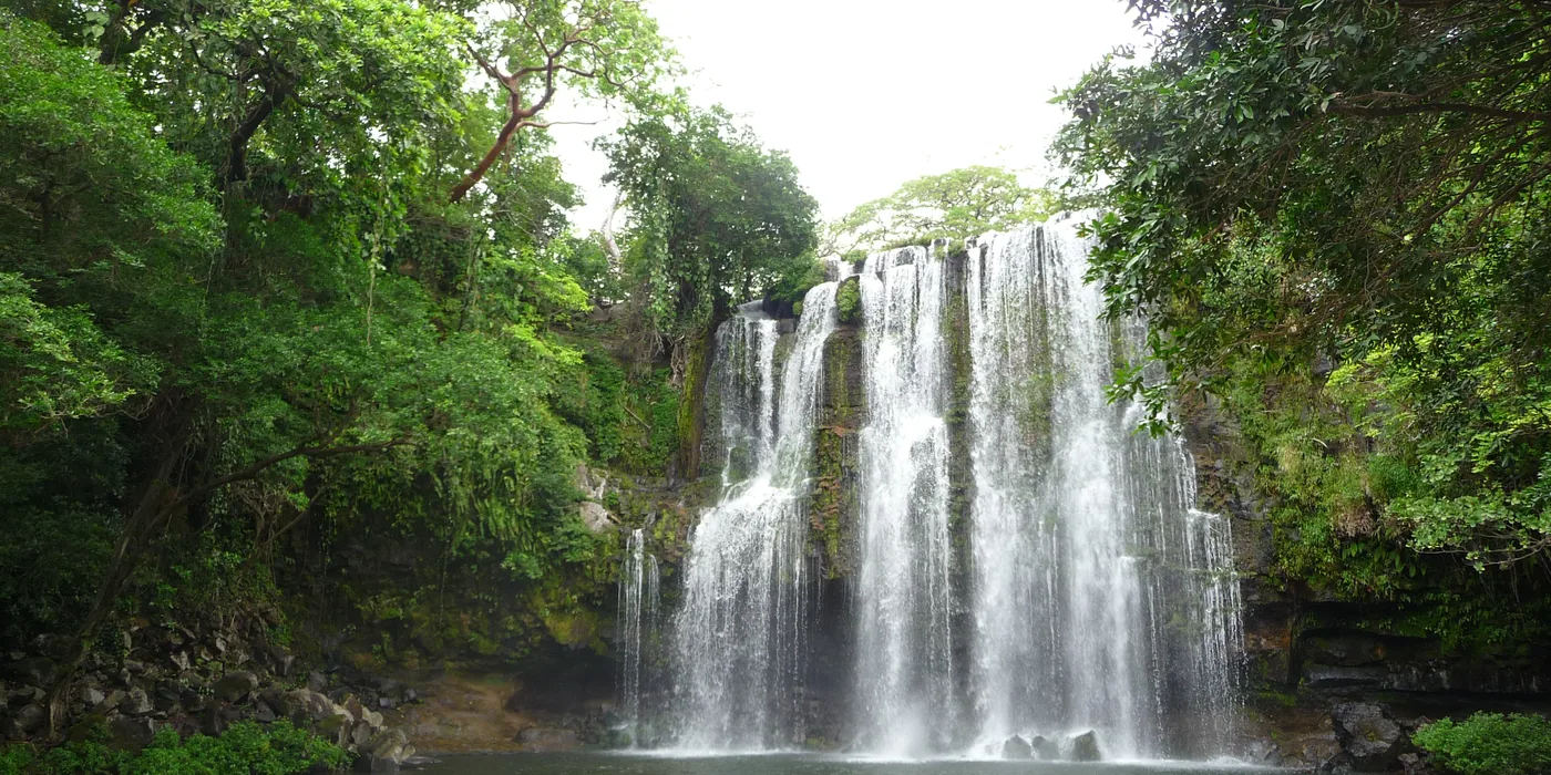 The wide curtain of Llanos de Cortés waterfall falling into a green pool, framed by dense forest on both sides