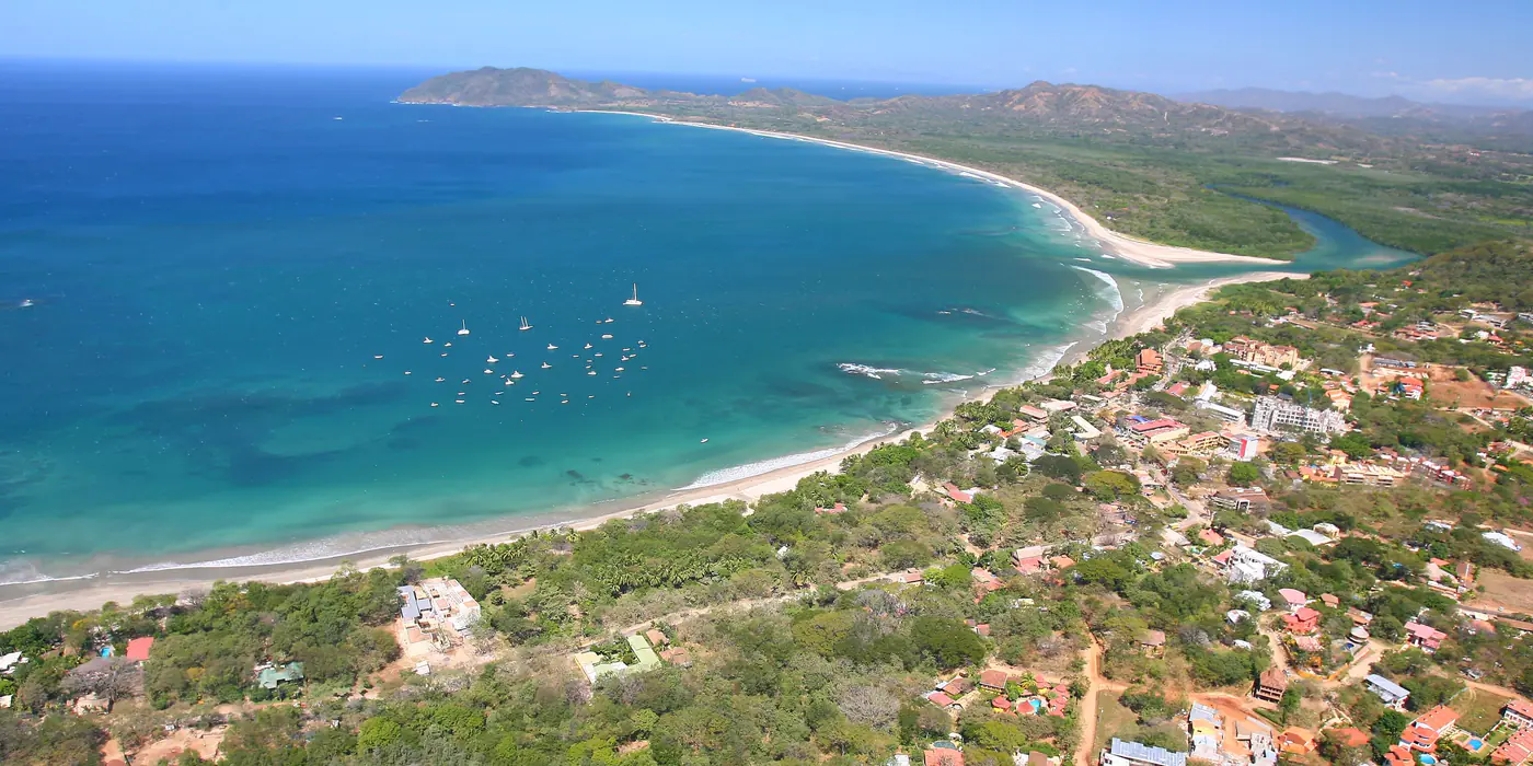 Aerial view of Tamarindo bay from the south: the town and beach in the foreground, boats moored in the bay, the mouth of the Tamarindo estuary and the long sweep of Playa Grande beyond it, with the Cabo Velas headland at the top