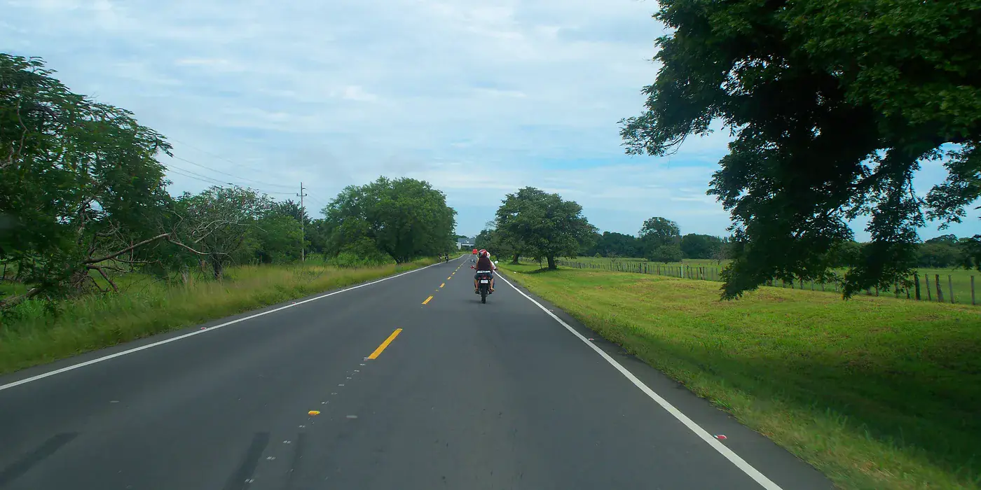 A two-lane paved stretch of the Pan-American Highway in Guanacaste seen through a windscreen, with fenced pasture and shade trees on both sides and a motorcyclist ahead