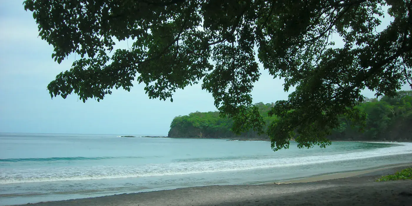 The Papagayo peninsula seen across calm water, low forested hills running down to a pale curving beach with almost no building visible