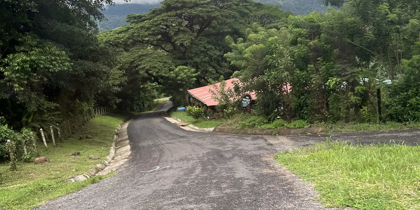 A narrow rural road in Guanacaste, Costa Rica, its worn asphalt running between a concrete drainage channel and an unpaved side turning, with a red-roofed building and forested hills beyond