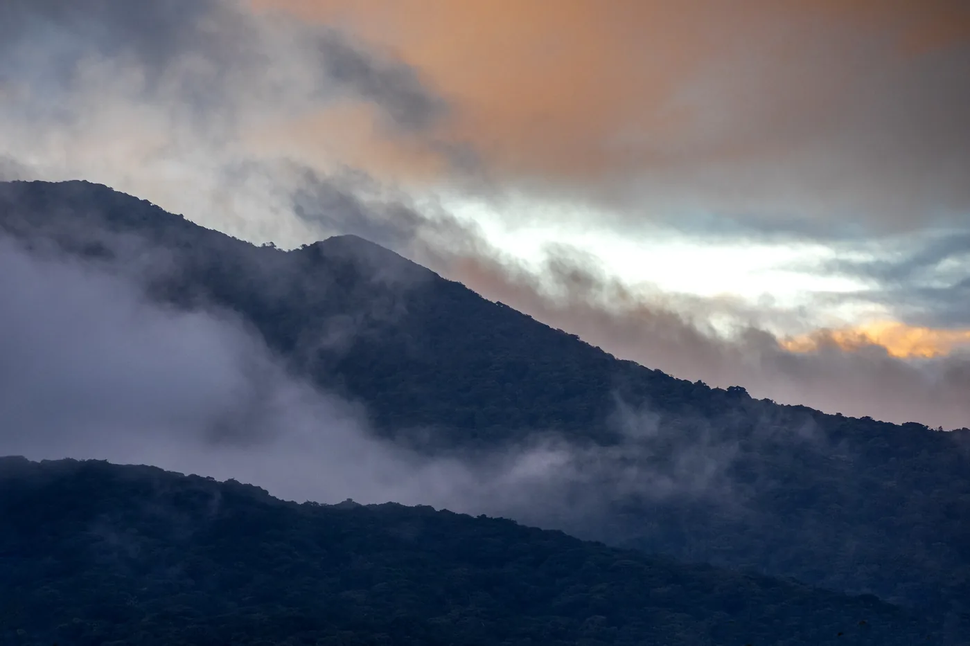 The forested flank of Rincón de la Vieja volcano at dusk, with cloud spilling over the ridgeline and an orange sky behind
