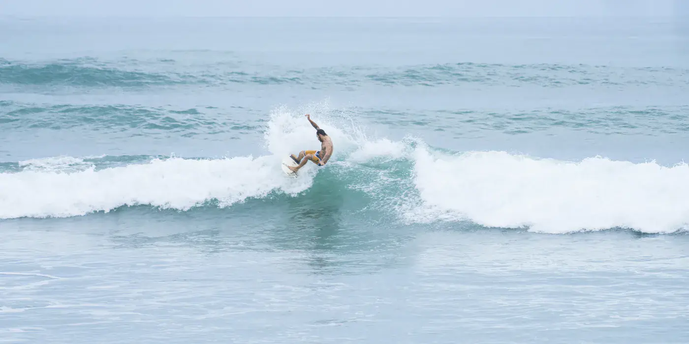 A surfer carving off the top of a chest-high wave at Playa Carmen on the Santa Teresa strip, with lines of whitewater behind and an overcast sky