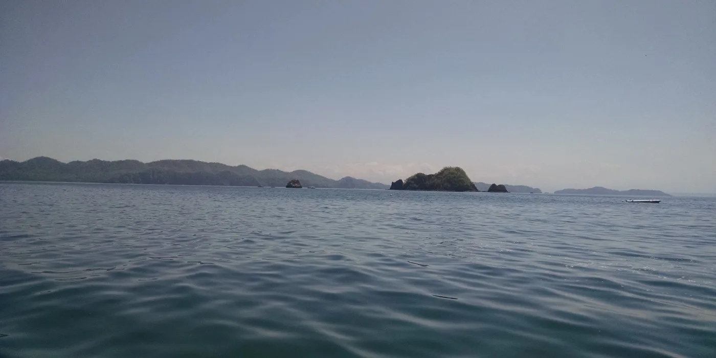 A rocky Pacific island dive and snorkeling site in Costa Rica, with clear shallow water breaking against dark volcanic rock under a bright sky