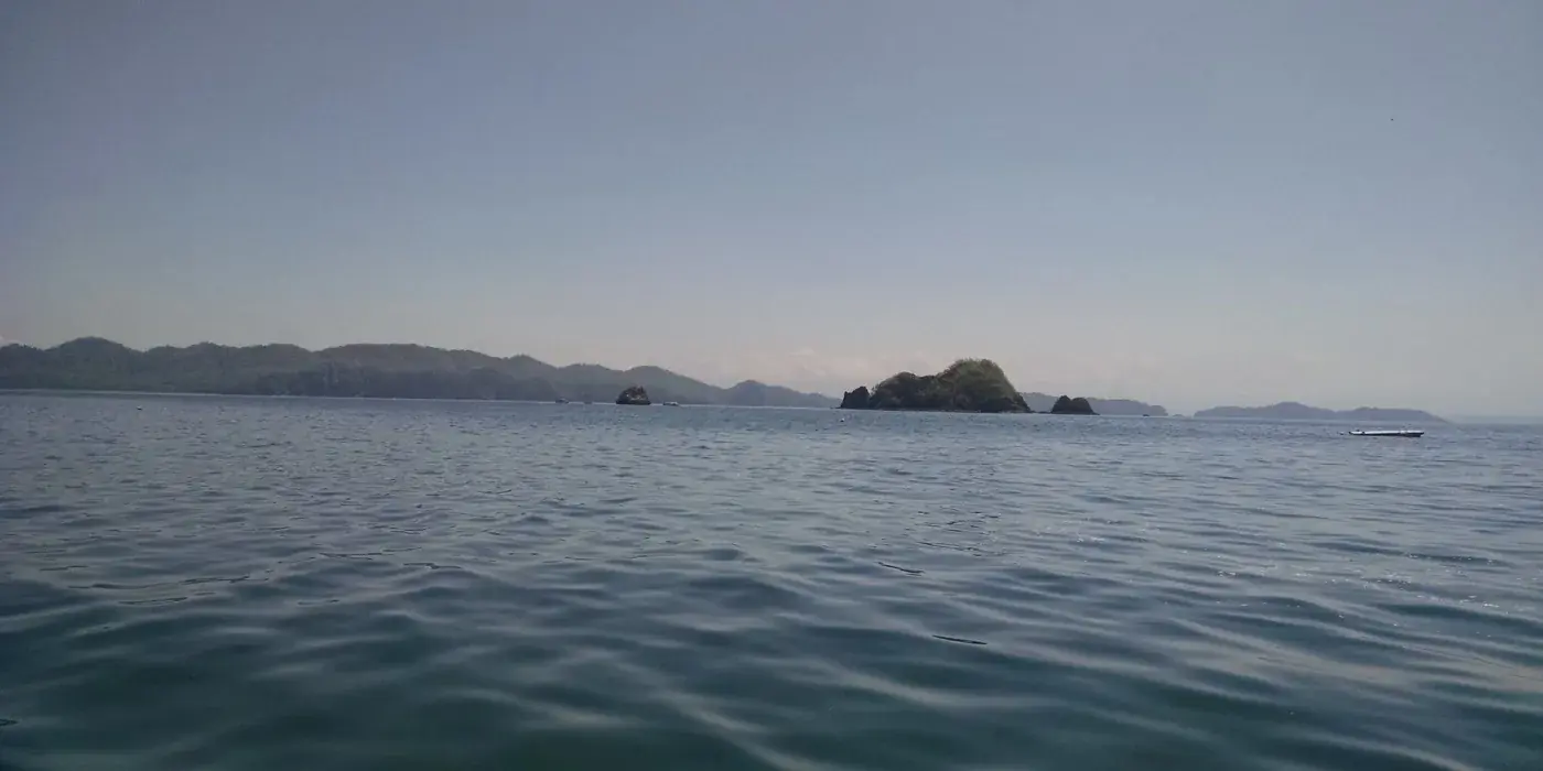 A rocky Pacific island dive and snorkeling site in Costa Rica, with clear shallow water breaking against dark volcanic rock under a bright sky