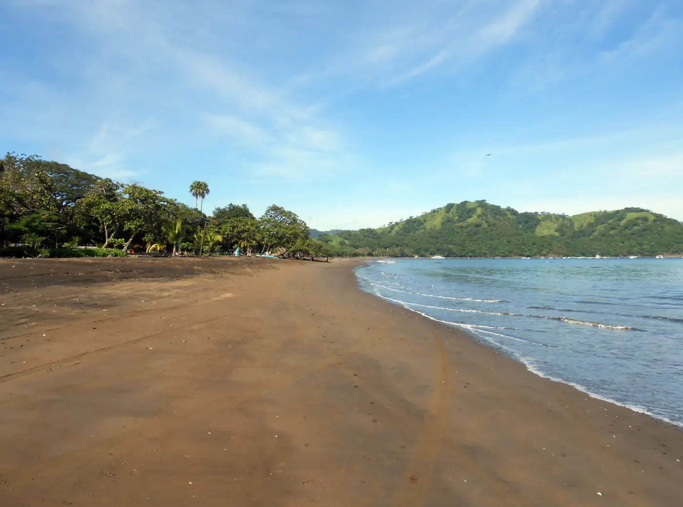 Dark-sand beach curving along the calm, sheltered bay at Playas del Coco, Guanacaste, with green hills and moored boats in the distance