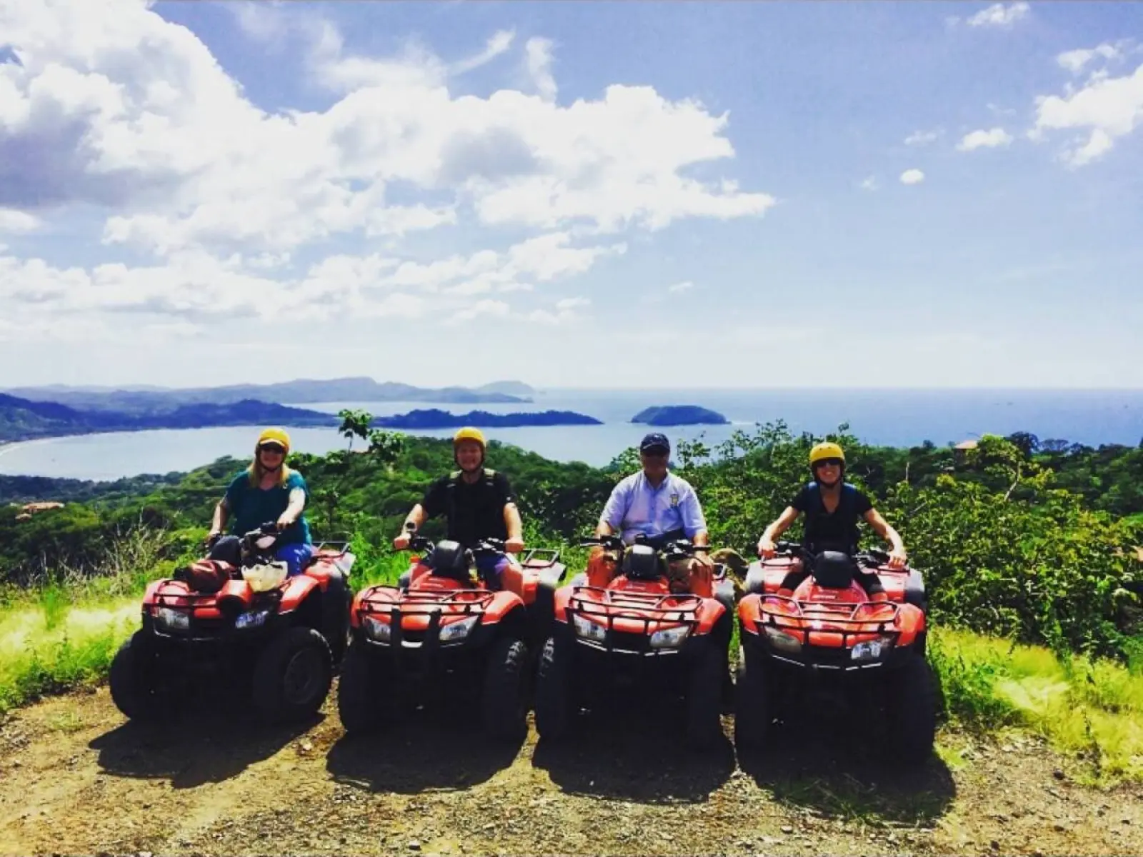 Four Flamingo Adventures riders on red ATVs at a hilltop lookout above a bay with islands and the Pacific Ocean behind