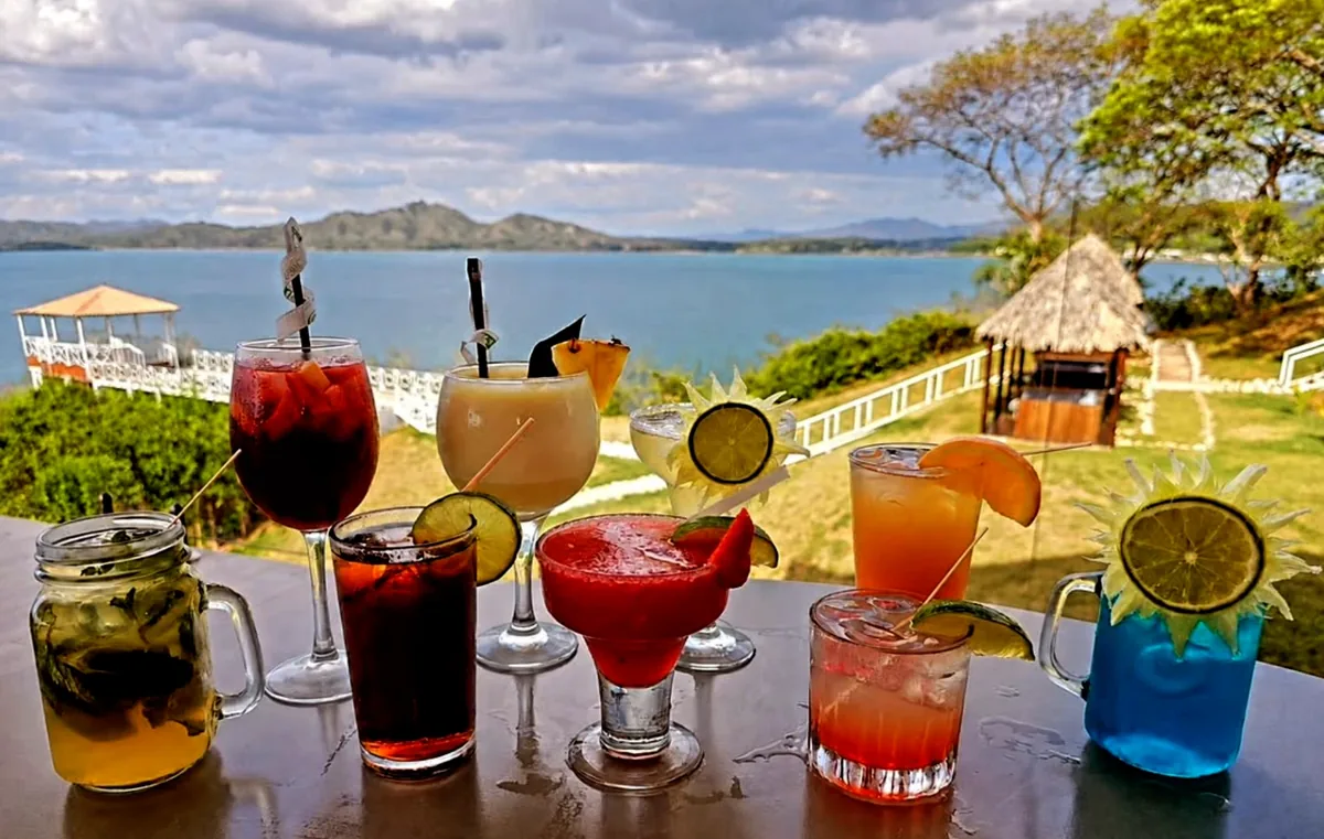 Eight cocktails lined along a rail above the bay, including sangria, pina colada, margarita and a blue drink garnished with lime