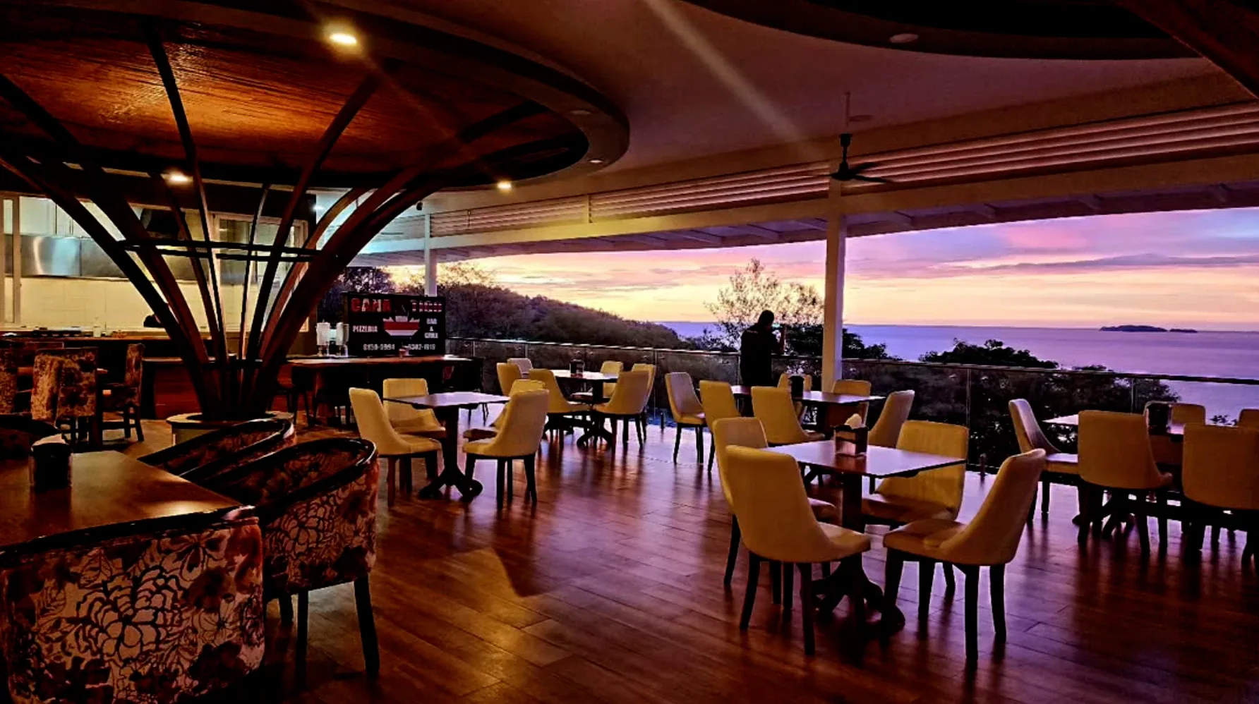 Open-air dining room at Cana Tico at sunset, tables and upholstered chairs under a curved timber canopy, looking out over the bay